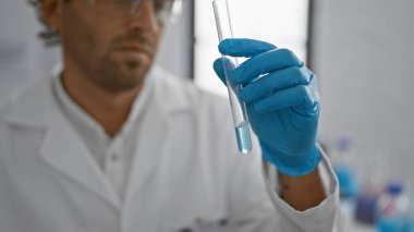 Sakallı İspanyol bir adam, kapalı bir laboratuvar ortamında bilim ve sağlık hizmetlerini temsil eden bir test tüpünü inceliyor..