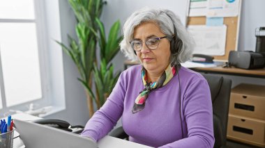 Kulaklıklı yaşlı bir kadın modern ofiste dizüstü bilgisayar kullanıyor..