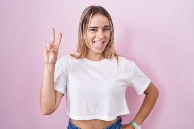 Young blonde woman standing over pink background smiling with happy face winking at the camera doing victory sign with fingers. number two. 