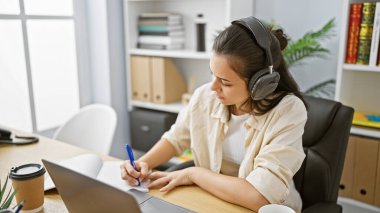Laptop ve kulaklık kullanan genç ve güzel İspanyol iş kadını ofiste not alıyor.