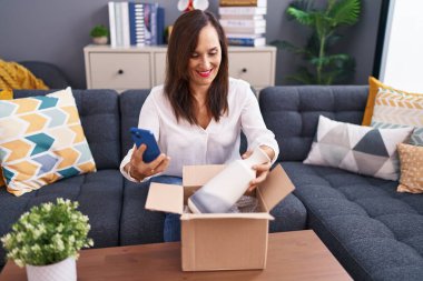 Genç, güzel, İspanyol bir kadın akıllı telefon kullanarak evdeki karton kutuları açıyor.