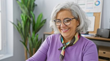 Ofiste gri saçlı, gözlüklü ve mor kazaklı, gülümseyen olgun bir kadın..