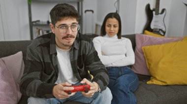 Kollarını kavuşturmuş üzgün bir kadın modern bir oturma odasında video oyunu oynayan odaklanmış bir adamın yanında oturuyor..