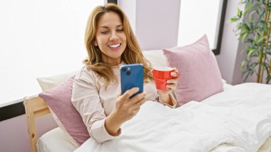 Gülümseyen İspanyol bir kadın, rahat bir yatak odasında akıllı telefonunu karıştırırken sabah kahvesini yatakta içer..
