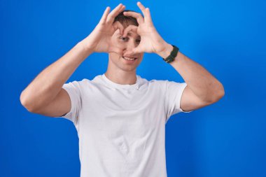 Beyaz tenli sarışın adam mavi arka planda duruyor. El ve parmaklarıyla kalp şeklinde. Tabelanın içinden bakıp gülümsüyor. 