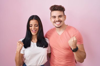 Pembe arka planda duran genç İspanyol çift mutlu ve heyecanlı bir şekilde kollarını kaldırıp, gülümseyip başarı için çığlık atıyorlar. kutlama konsepti. 