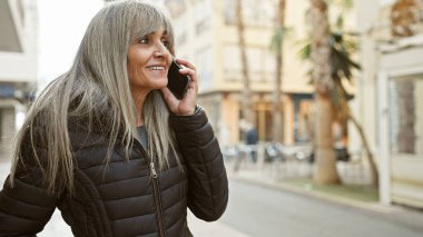 Gri saçlı, gülümseyen olgun bir kadın palmiye ağaçlarıyla dolu bir caddede dikilirken cep telefonuyla konuşuyor..