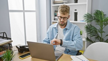 Sakallı ve mavi gözlü genç beyaz adam modern ofis ortamında zamanı kontrol ediyor.