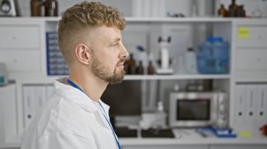 Beyaz, sakallı ve mavi gözlü bir adam laboratuvar ortamında laboratuvar önlüğü giyiyor..