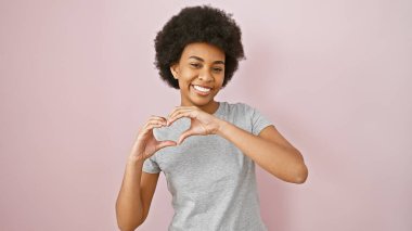 Gülümseyen Afro-Amerikan kadını ellerini pembe bir arka plana dayayarak bir kalp oluşturuyor, pozitiflik sergiliyor.