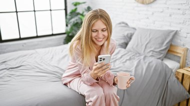 Pijama giyen, elinde kahve fincanı tutan, rahat bir yatak odasında akıllı telefon kullanan, gülümseyen beyaz bir kadın..
