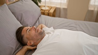 Çekici genç İspanyol adam rahatça rahatlar, elleri başının üstünde dinlenir, güneşli bir yatak odasında rahat bir yatakta uzanır, sabah rahatlığını ifade eder..