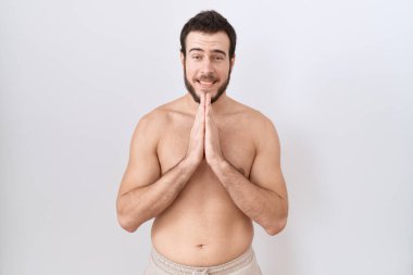 Beyaz arka plan üzerinde üstsüz duran genç İspanyol adam elleri ile birlikte dua ederek af diliyor kendinden emin bir şekilde gülümseyerek.. 