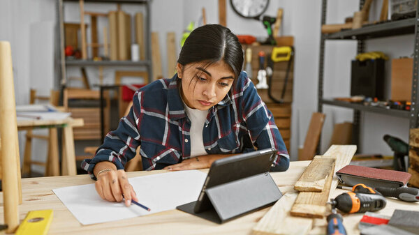 Индейка рисует планы в столярной мастерской, окруженной деревом и инструментами.