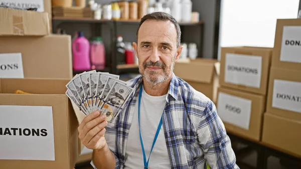 Community service hero, middle age man with grey hair volunteers at local charity center, holding a wealth of dollars for donation