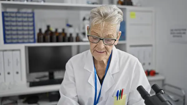 Grey-haired senior woman scientist wearing glasses, engrossed in crucial medicine research lab work