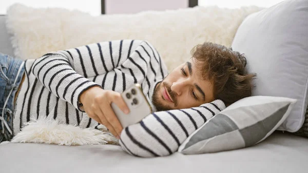 Handsome young arab man, with a serious expression, deep into online texting, lying relaxed on his living room sofa at home, using his smartphone technology indoors.