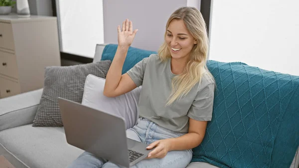 Smiling woman using laptop on couch, waving during a video call at home.