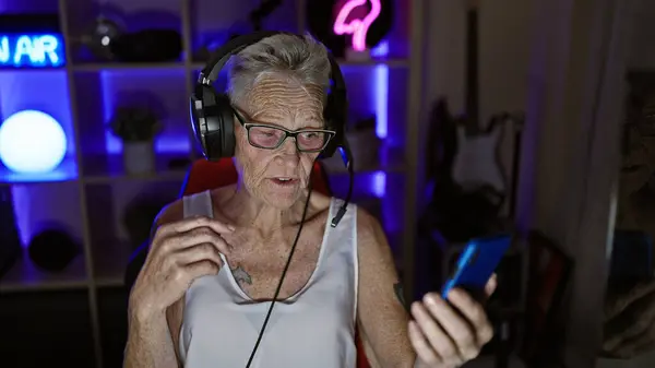 Tech-savvy, grey-haired senior woman enthrals gaming world as she streams live video call, gamepad in hand, from her darkly lit gaming room - technology, entertainment and age have no bar!