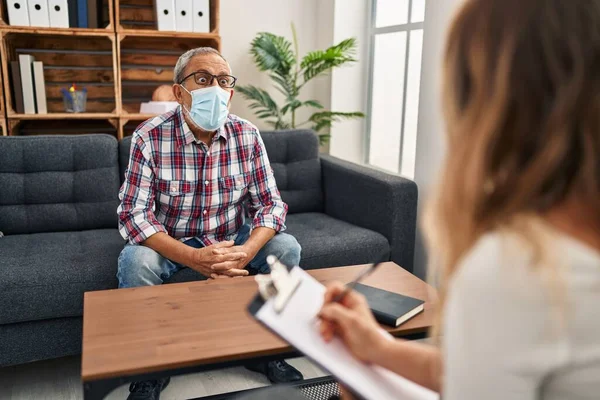 Korkmuş kıdemli bir adam, yüzü inançsızlıkla dolu, psikoloji kliniğinde şaşkın bir şekilde oturuyor, tıbbi maske destansı bir sürprizle ağzı açık.