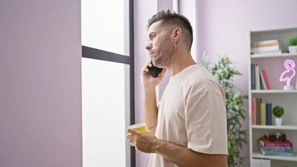 Serious young hispanic man in a deep conversation over the phone, throwing sad looks through the window while sipping coffee at home in the morning.