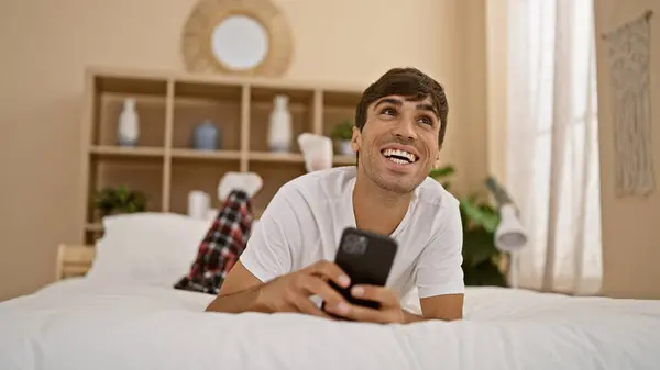 Attractive, young hispanic man enjoying a laid-back morning, comfortably lying in bed, happily texting on smartphone with a gleaming smile in a cosy bedroom home interior