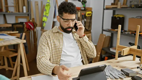 Young hispanic man with beard multitasking on a phone call in a carpentry workshop.