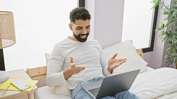 Smiling bearded man using laptop and gesticulating in a bright bedroom.