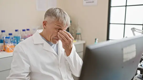 一位身穿实验室外套的疲惫的老人在明亮的实验室里与计算机一起工作时捏住了鼻梁 — 图库照片