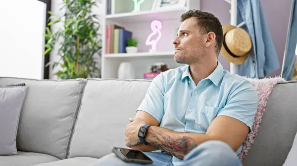 Sakallı ve dövmeli yakışıklı bir İspanyol modern bir oturma odasında düşünceli bir şekilde oturuyor..