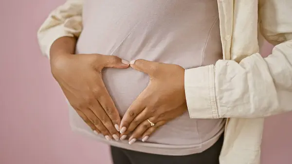 期待着穿着休闲装的年轻女子 站在孤立的粉红墙上 双手捧着心形的符号来到怀孕的腹部 表达着可爱的母性情感 — 图库照片