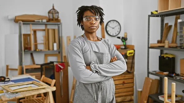 Confident woman with dreadlocks standing arms crossed in a well-equipped carpentry workshop.