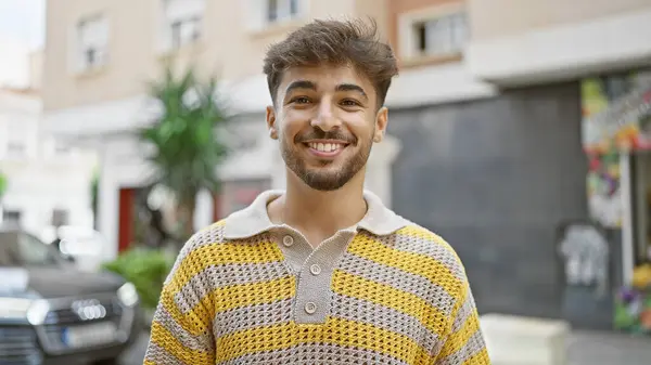 Confident young arab man, cheerfully enjoying his time outdoors standing on city street, his bearded face expressing pure joy and happiness through a handsome smile