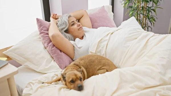 Mature woman resting with dog in a bright bedroom, reflecting a serene, comfortable indoor lifestyle.