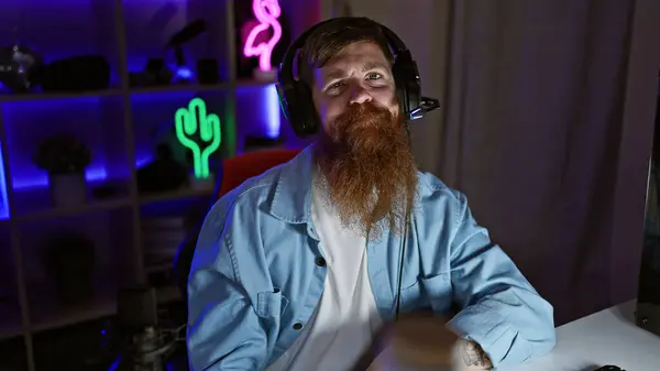 Staunch young redhead man streamer, seriously immersed in digital gaming world, donning headphones in the dimly lit gaming room