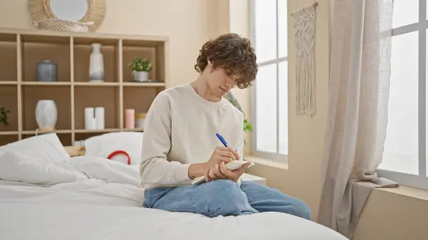 A young man with curly hair and tattoo writes in a notebook while sitting comfortably on a white bed in a sunlit bedroom.