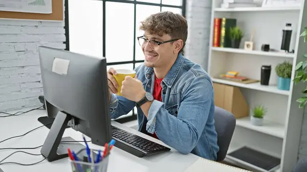 Confident young hispanic man at work, smiling business professional using computer, drinking morning coffee in the elegant office interior