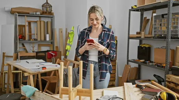 Stock image A young woman smiles while using her phone in a cluttered woodworking workshop.