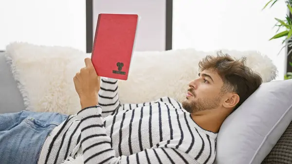 Young arab man reading book lying on sofa at home