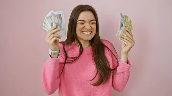 Happy young woman holds peruvian soles with a wink against a pink wall, showing wealth and joy.