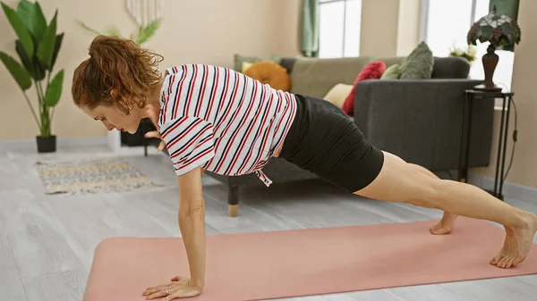 A focused woman practices a plank exercise indoors on a pink yoga mat within a cozy living room setting.