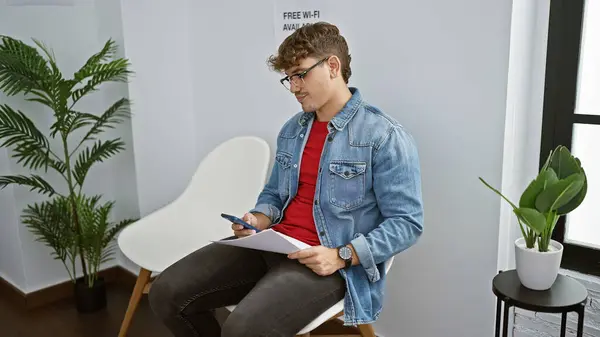 Handsome, young hispanic man engrossed in a serious text conversation on his smartphone, while anxiously clutching documents and sitting in a waiting room chair, indoor portrait.