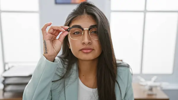 stock image A professional south asian woman with long hair and glasses poses confidently in a bright office setting.