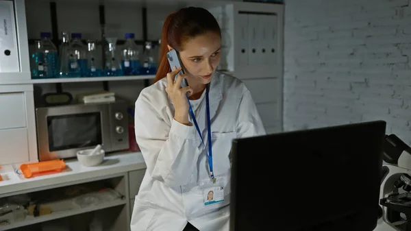 A focused woman scientist multitasking in a lab with a phone call and computer work, amidst lab equipment and white brick backdrop.