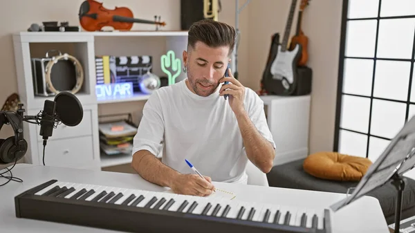 stock image Young hispanic male muso, pianist, in his element, writing notes in a notebook, engaged in a serious phone conversation inside a buzzing music studio