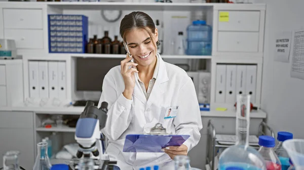 Stunning young hispanic woman scientist, reading report and talking on smartphone at lab work table, mastering the art of chemistry research