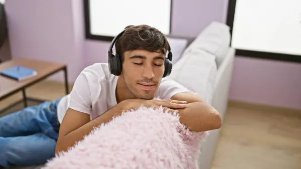 Portrait of a relaxed, confident young hispanic man, joyfully enjoying the sound of music, listening through headphones, sitting indoors on the sofa in a cosy, modern apartment.