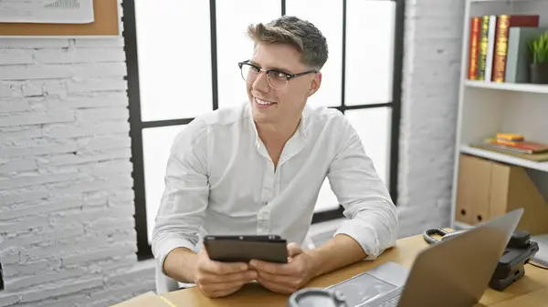Confident young caucasian man enjoying success as he works on laptop, utilizing touchpad in indoor office setting, exuding professional business charm from behind glasses at his desk.