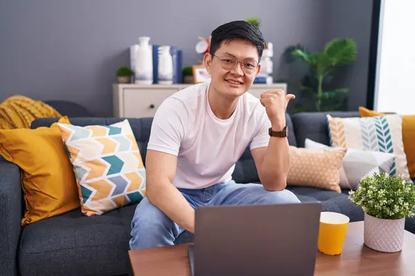 Genç Asyalı adam evinde dizüstü bilgisayar kullanıyor. Koltukta oturuyor. Arka tarafı işaret ediyor. 