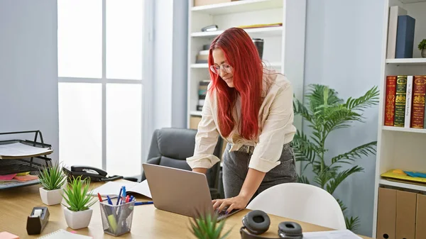 Confident young redhead business woman joyfully working online at her office, managing success with laptop, beautiful irish employee smiling behind glasses at her workplace.
